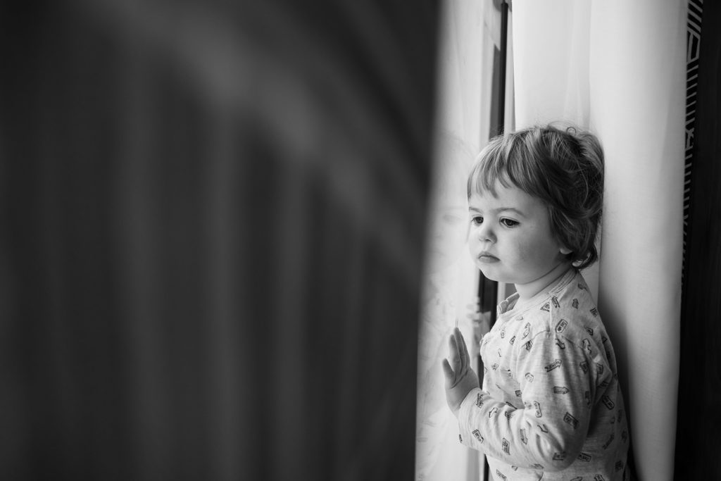 Reportage du quotidien près de Rennes. Portrait noir et blanc d'un jeune enfant regardant par une baie vitrée d'un air rêveur.