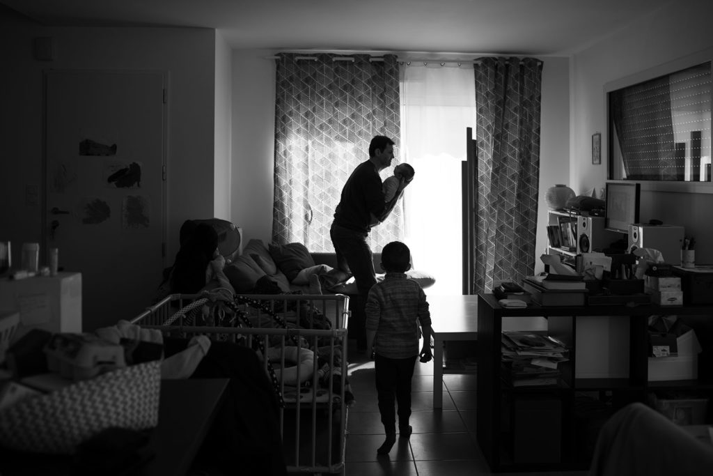 Reportage du quotidien près de Rennes. Photographie en noir et blanc d'un papa portant son bébé en contre-jour.